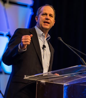 Man in dark suite speaking from a lectern on a stage with a dark blue background.