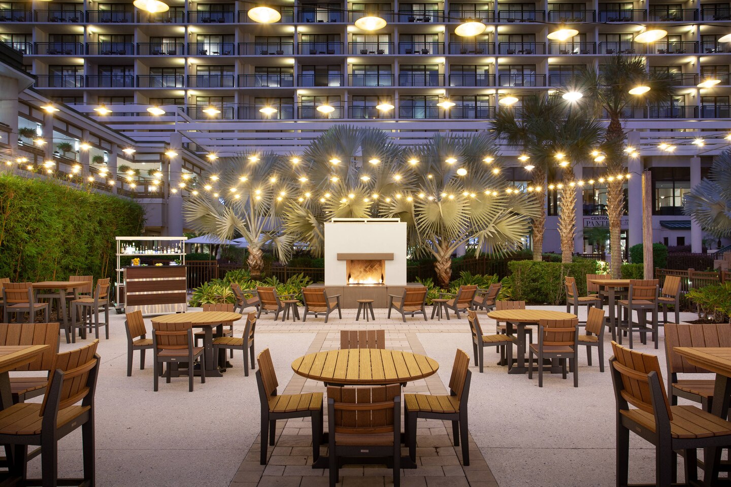 A patio with wooden chairs and black accent lit up with string lights over head outside with palm trees and a hotel in the background
